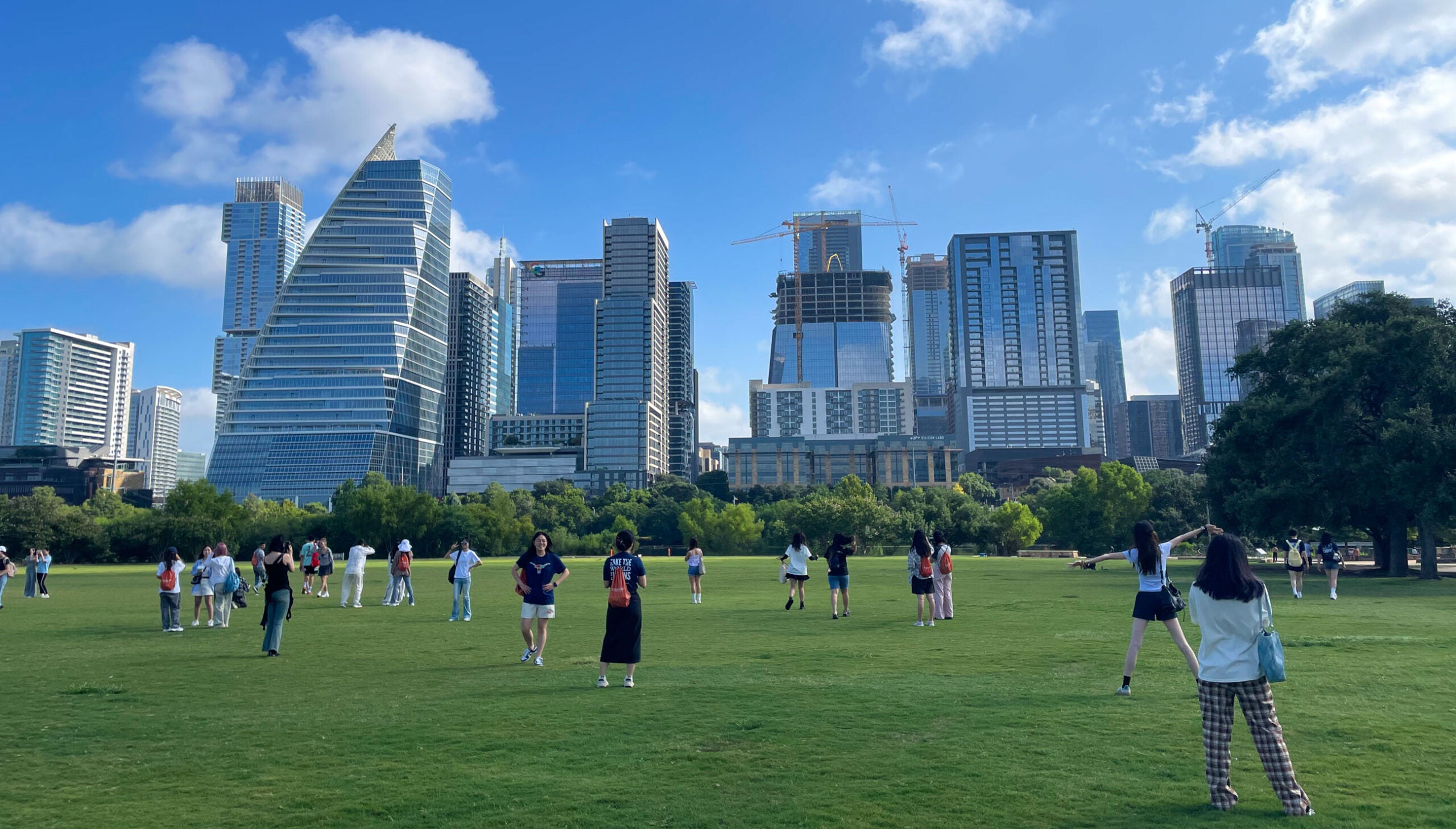 People gather in a park filled with green grass with a beautiful view of the Austin City Skyline in the background on a blue sky day