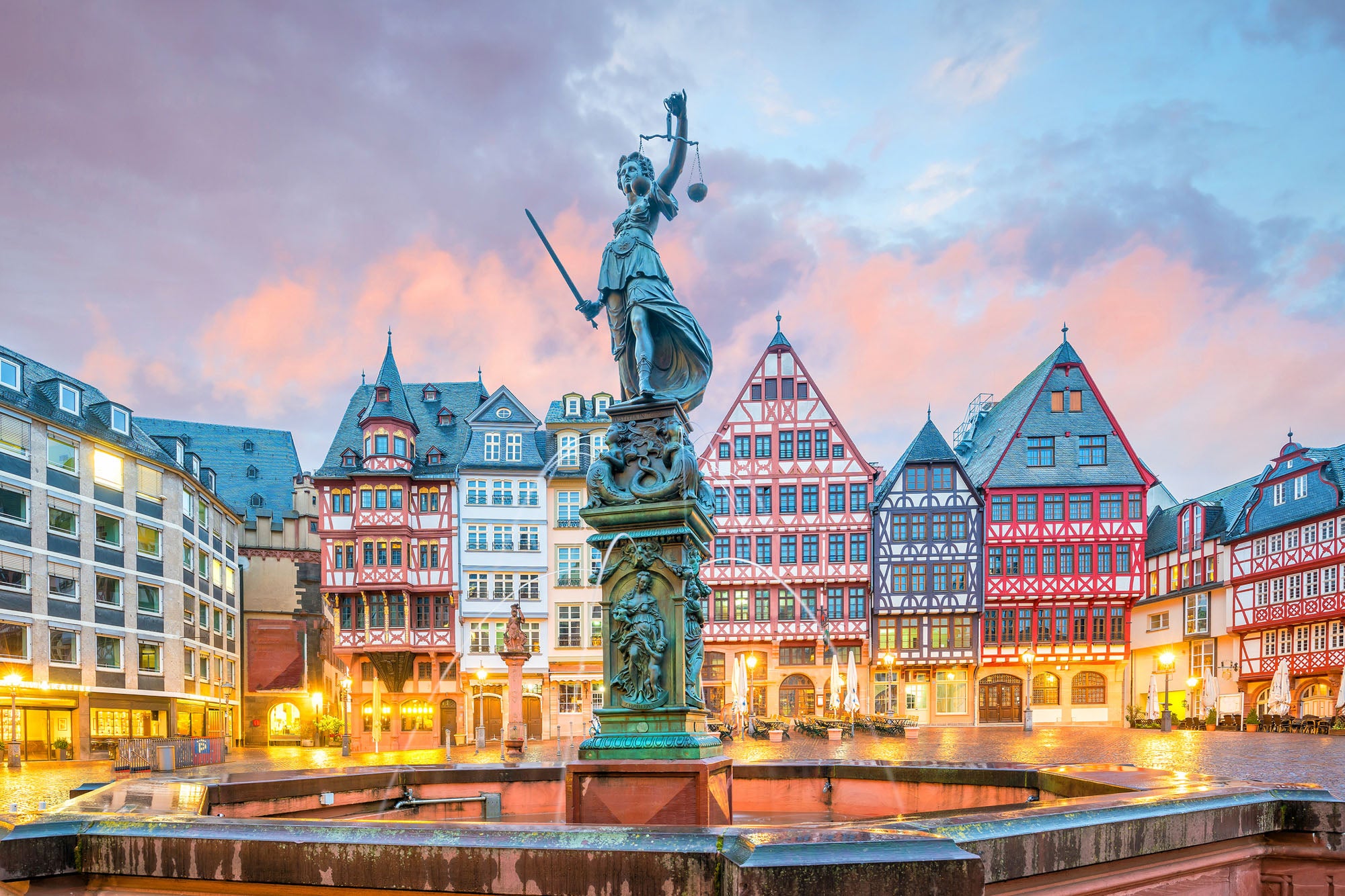 A large bronze statue known as the Altstadt and fountain stand at the center of Römerberg, a historic town square in Frankfurt, Germany at twilight