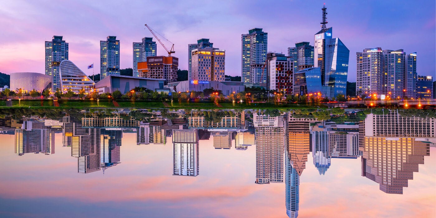 A composite image of the city skylines of Austin, TX and Daejeon, South Korea perfectly reflected in their respective rivers that run beside them during a beautiful purple, blue, and pink sunset.