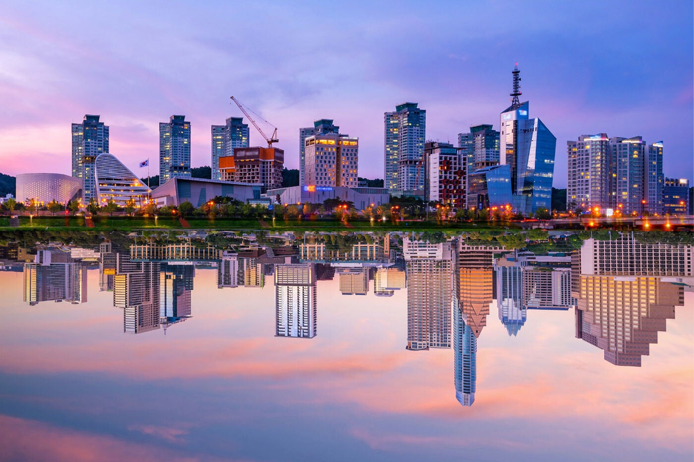 A composite image of the city skylines of Austin, TX and Daejeon, South Korea perfectly reflected in their respective rivers that run beside them during a beautiful purple, blue, and pink sunset.