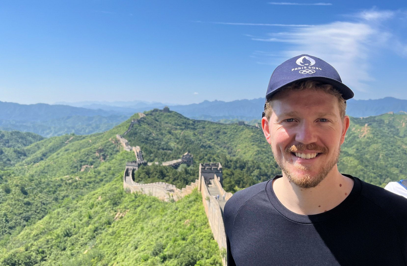 Sebastian DeBeurs smiles while standing atop the Great Wall of China on a beautiful blue sky day